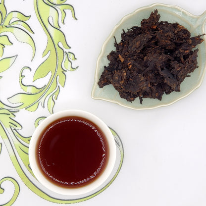 Dried tea leaves on a small plate with a cup of brewed tea on a decorative white and green background.