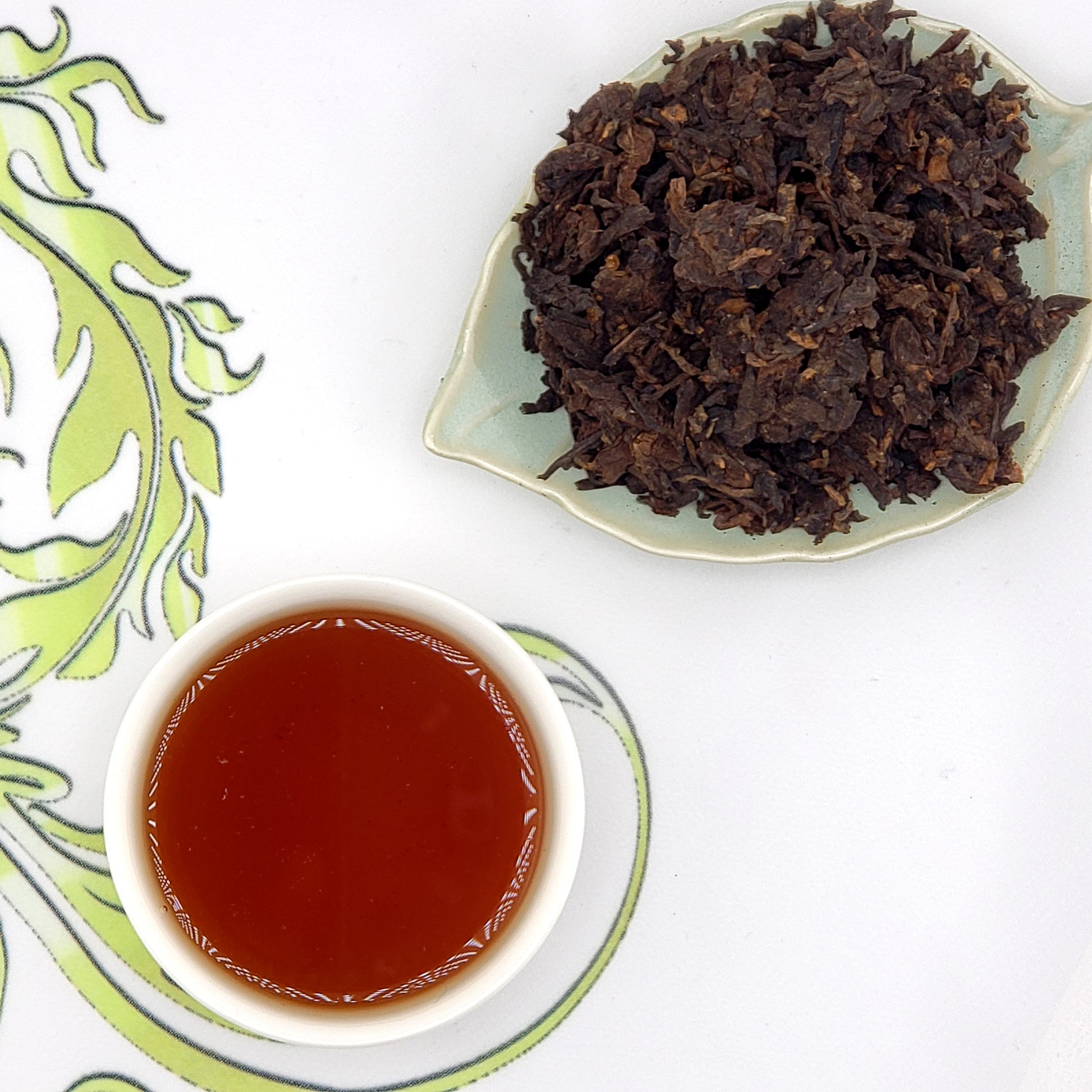Dried tea leaves on a leaf-shaped plate with a cup of brewed tea on a decorative mat.