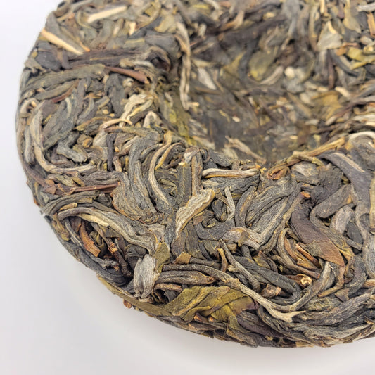 A close-up of a circular compressed tea cake made of tightly packed dried tea leaves, showing varied shades of dark gray, olive green, and brown on a plain white background.