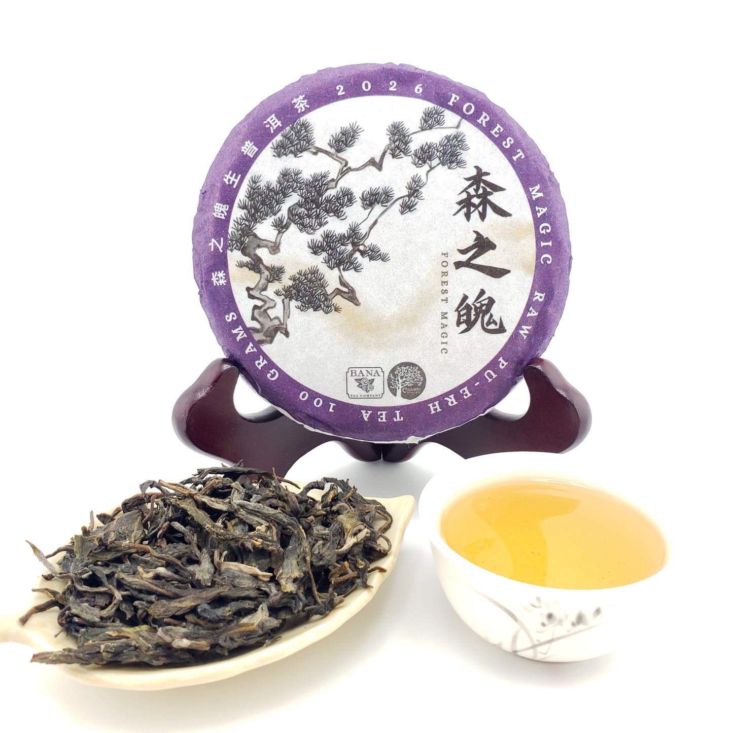 A round tea disc package on a small wooden stand, with loose dark tea leaves in a small dish and a white cup of pale golden tea in front, all on a white background. The package label reads 2026 Forest Magic Pu Erh Tea.