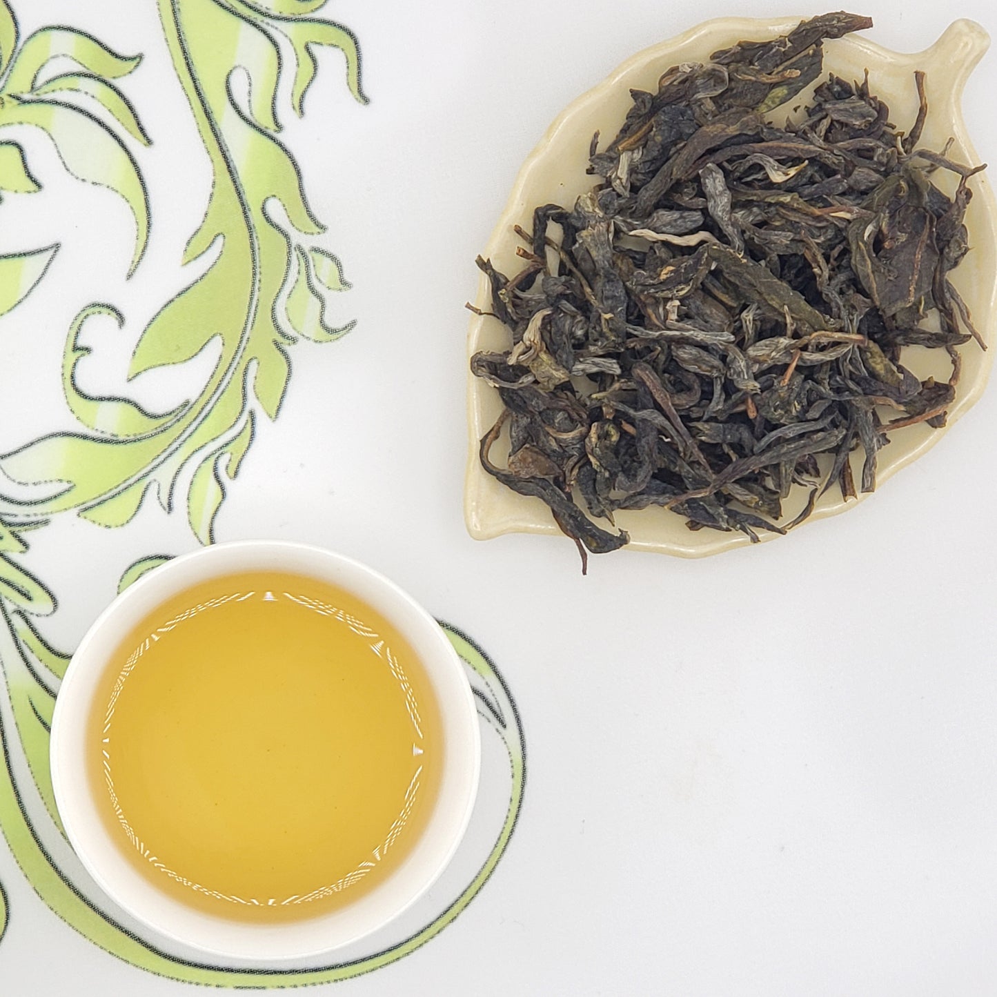 A cup of pale golden tea on a white saucer next to a leaf-shaped dish filled with loose dark tea leaves, arranged on a white surface with a green patterned cloth.