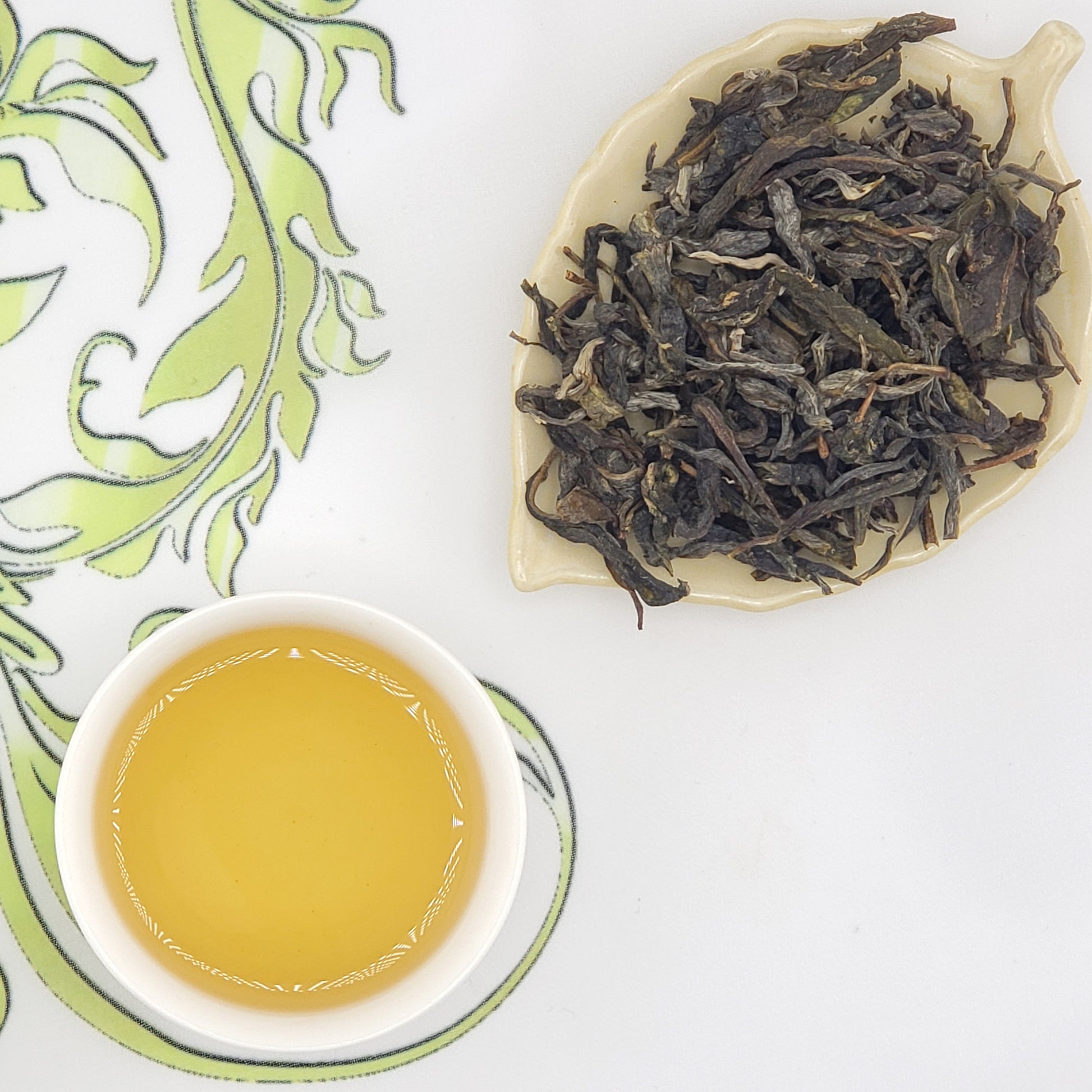 A cup of pale golden tea on a white saucer next to a leaf-shaped dish filled with loose dark tea leaves, arranged on a white surface with a green patterned cloth.