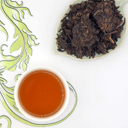 Tea cup with brewed tea and a dish of dry tea leaves on a decorative mat.