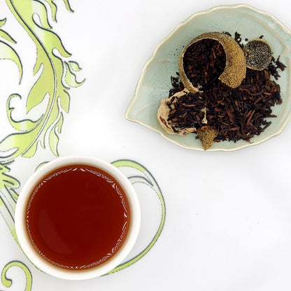 Tea leaves spilling out of a broken green mandarin shell on a plate next to a cup of tea on a decorative surface