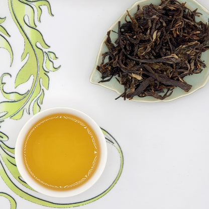 Dried tea leaves on a plate with a cup of brewed tea on a decorative mat.