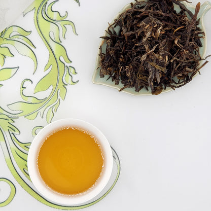 Dried tea leaves on a plate with a cup of brewed tea on a decorative white and green background.