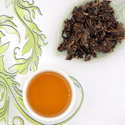 Dried tea leaves on a plate with a cup of brewed tea on a decorative white and green background.