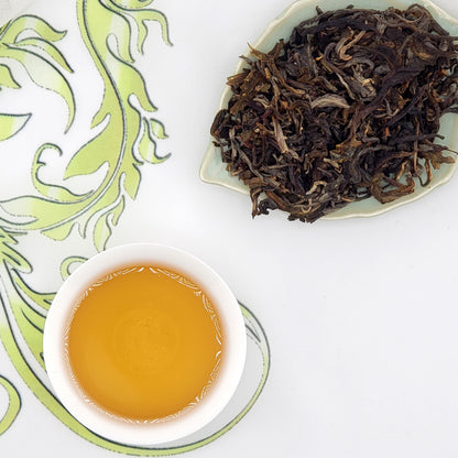 Dried tea leaves on a white plate with a cup of brewed tea on a decorative green and white background.