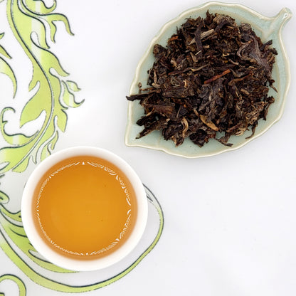 Dried tea leaves on a leaf-shaped plate with a cup of brewed tea on a decorative white and green background.