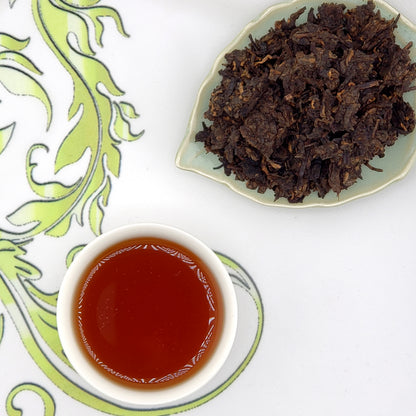 Dried tea leaves on a plate with a cup of brewed tea on a decorative mat.