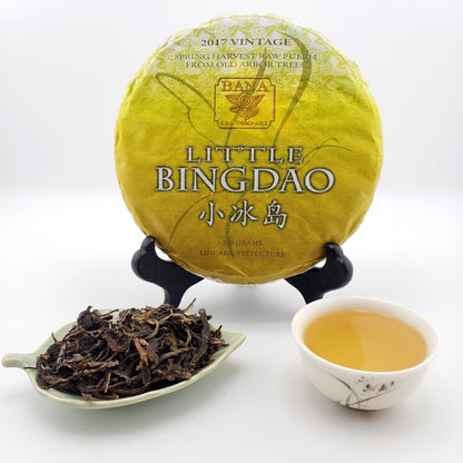Yellow tea cake labeled 'Little Bingdao' with a teacup of tea and a scoop of tea leaves on a white background.