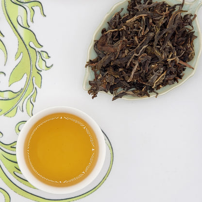 Dried tea leaves on a plate with a cup of brewed tea on a decorative white and green background.