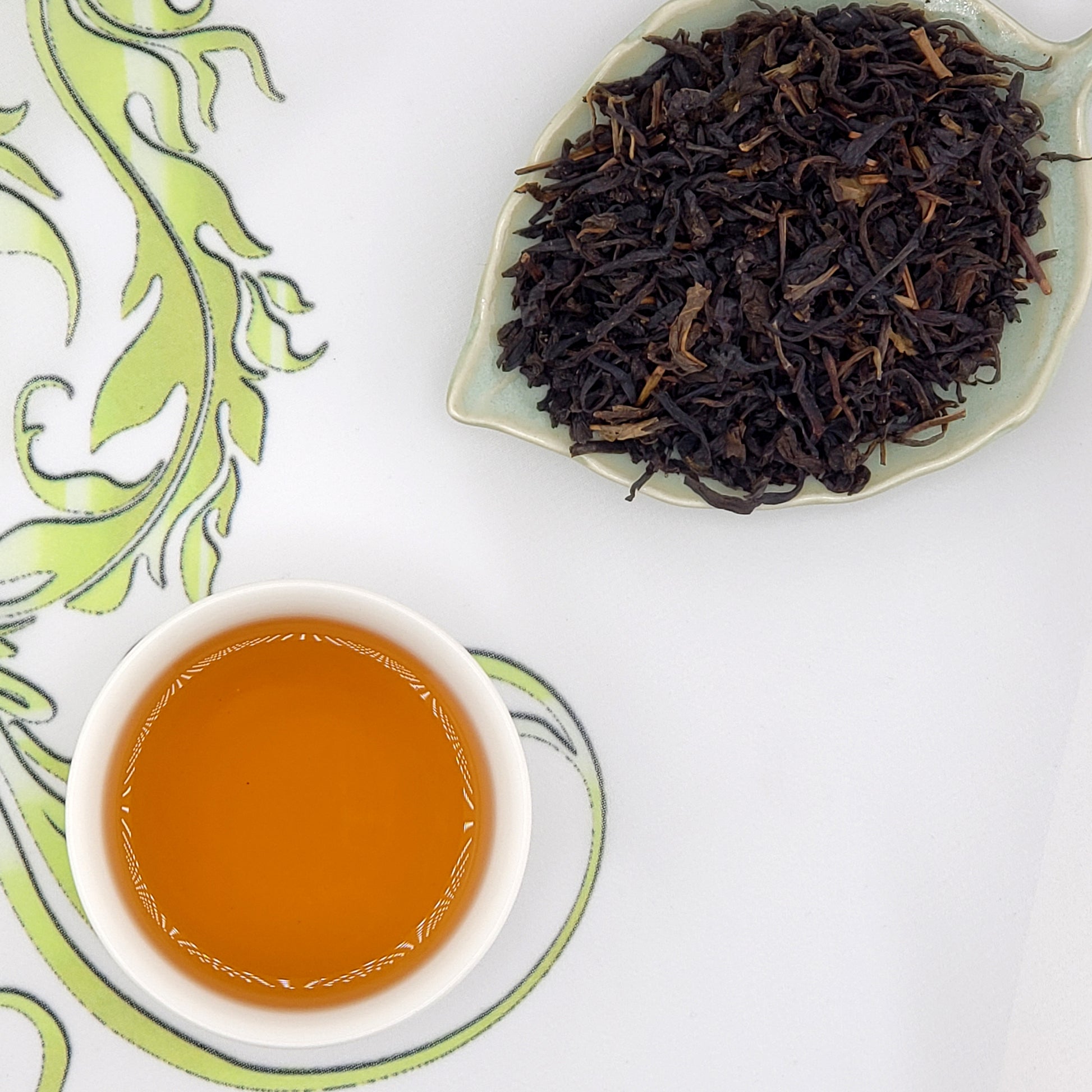 Dry black tea leaves on a leaf-shaped plate with a cup of brewed tea on a decorative white surface.