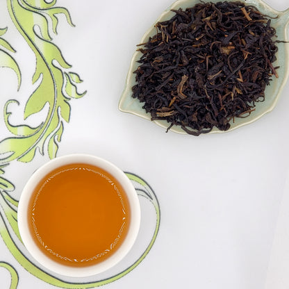 Dry black tea leaves on a leaf-shaped plate with a cup of brewed tea on a decorative white surface.