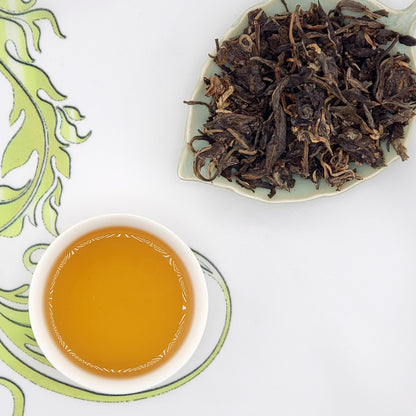Dried tea leaves on a plate next to a cup of brewed tea on a decorative mat.
