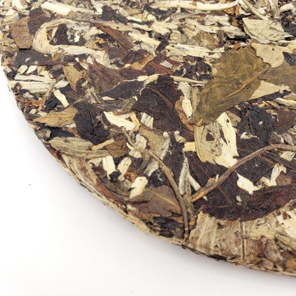 A close-up of a compressed cake of loose-leaf tea, showing mottled dark brown, tan, and off-white dried leaves and bark pieces, photographed on a white background.