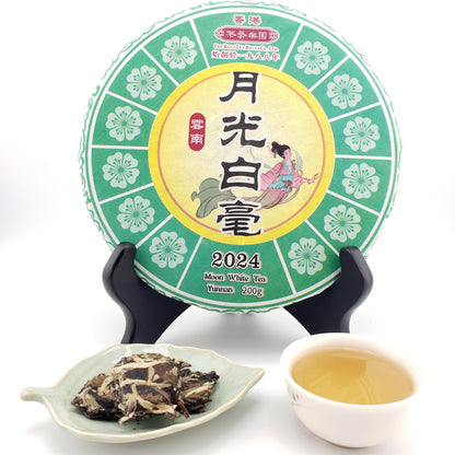 A round cake of Moonlight White Tea (Yunnan 2024) displayed on a black stand, with a small dish of loose dried tea leaves and a cup of brewed tea in front, set against a clean white background.
