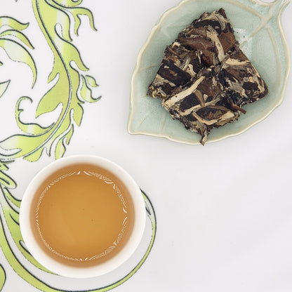 A top-down view of a white teacup filled with amber tea and a leaf-shaped dish holding dark compressed tea blocks, set on a white surface with a green patterned cloth.