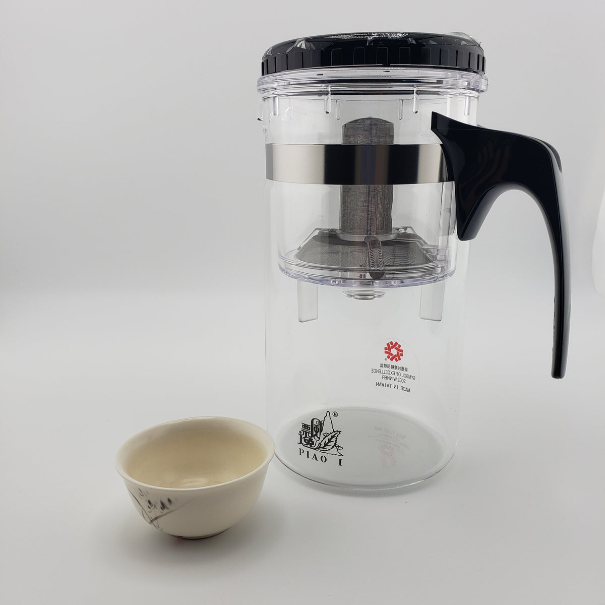 A clear glass tea infuser pitcher with a black lid, black handle, and stainless steel infuser basket, shown next to a small ceramic teacup on a light gray background. The brand name 'PIAO I' is printed on the bottom of the pitcher.