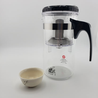 A clear glass tea infuser pitcher with a black lid, black handle, and stainless steel infuser basket, shown next to a small ceramic teacup on a light gray background. The brand name 'PIAO I' is printed on the bottom of the pitcher.