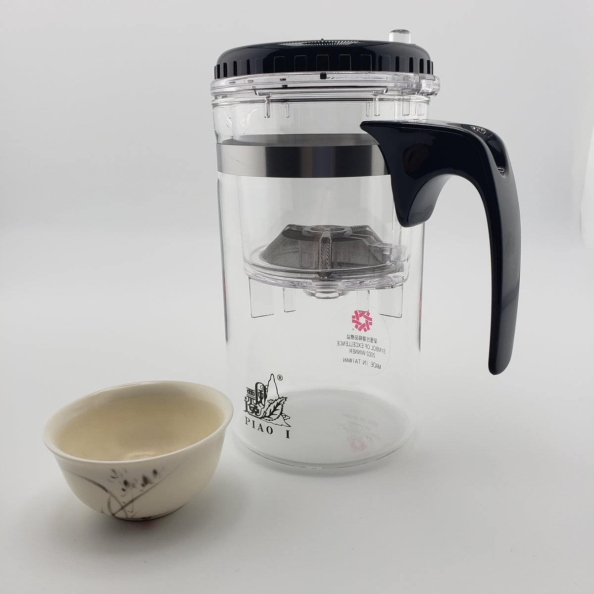 A clear glass teapot with a black lid, black handle, and metal infuser, shown next to a small beige ceramic cup on a light gray background.