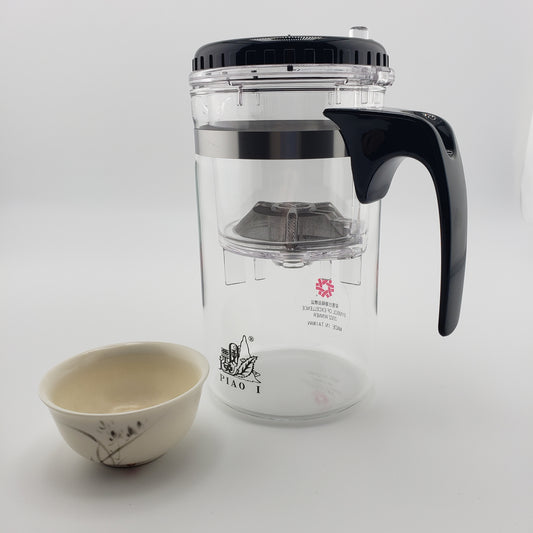 A clear glass teapot with a black lid, black handle, and metal infuser, shown next to a small beige ceramic cup on a light gray background.