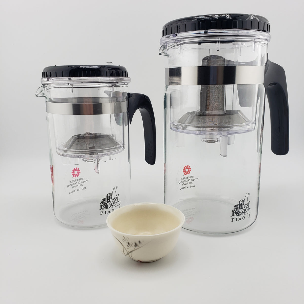 Two clear glass tea infusers with black lids and handles, shown on a white background with a small cream-colored ceramic teacup in front. The brand name 'PIAO I' is visible on the glass.