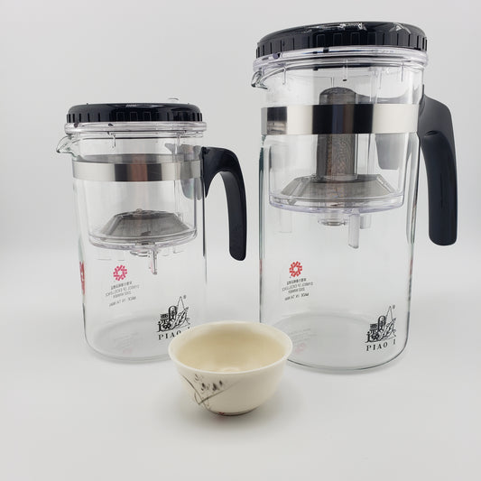 Two clear glass tea infusers with black lids and handles, shown on a white background with a small cream-colored ceramic teacup in front. The brand name 'PIAO I' is visible on the glass.