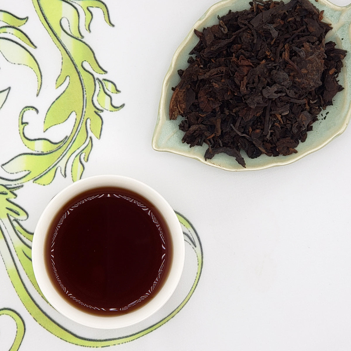 A cup of dark brewed tea on a white saucer next to a dish of loose dark tea leaves, arranged on a white surface with a green decorative pattern.