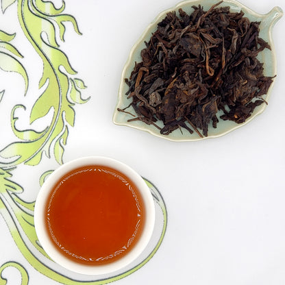Dried tea leaves on a leaf-shaped dish with a cup of brewed tea on a decorative mat.