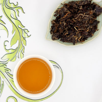 Dried tea leaves on a leaf-shaped plate with a cup of brewed tea on a decorative white surface.