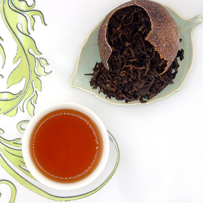 Tea leaves spilling out of a broken red mandarin shell and a cup of tea on a decorative plate with a floral pattern.