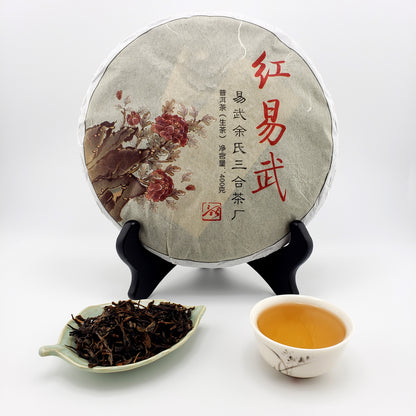 Tea cake with decorative wrapper on a stand, plate of dried tea leaves, and a cup of brewed tea.