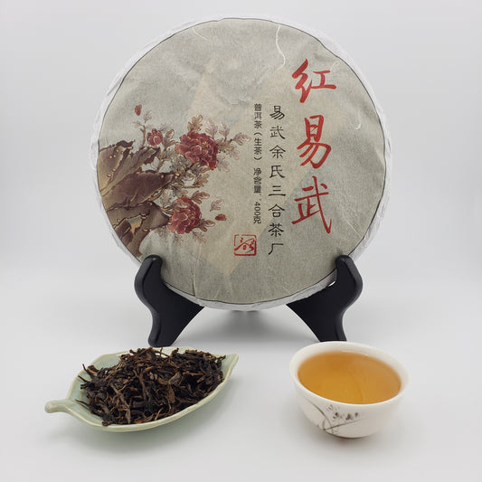 Tea cake with decorative wrapper on a stand, plate of dried tea leaves, and a cup of brewed tea.