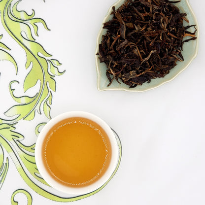 Dried tea leaves on a plate next to a cup of brewed tea on a decorative white and green background.