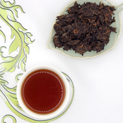 Dried tea leaves on a leaf-shaped plate with a cup of brewed tea on a decorative mat.