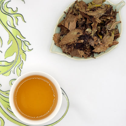 Dried tea leaves on a leaf-shaped dish with brewed tea in a small cup on a decorative white and green plate.