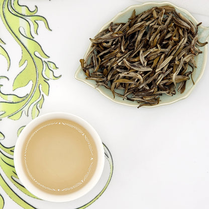 Dried tea leaves on a ceramic dish next to a cup of brewed tea on a decorative tablecloth.