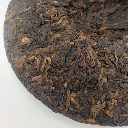 A close-up of a round compressed Pu-erh tea cake, showing the textured, dark brown and black leafy surface against a plain white background.