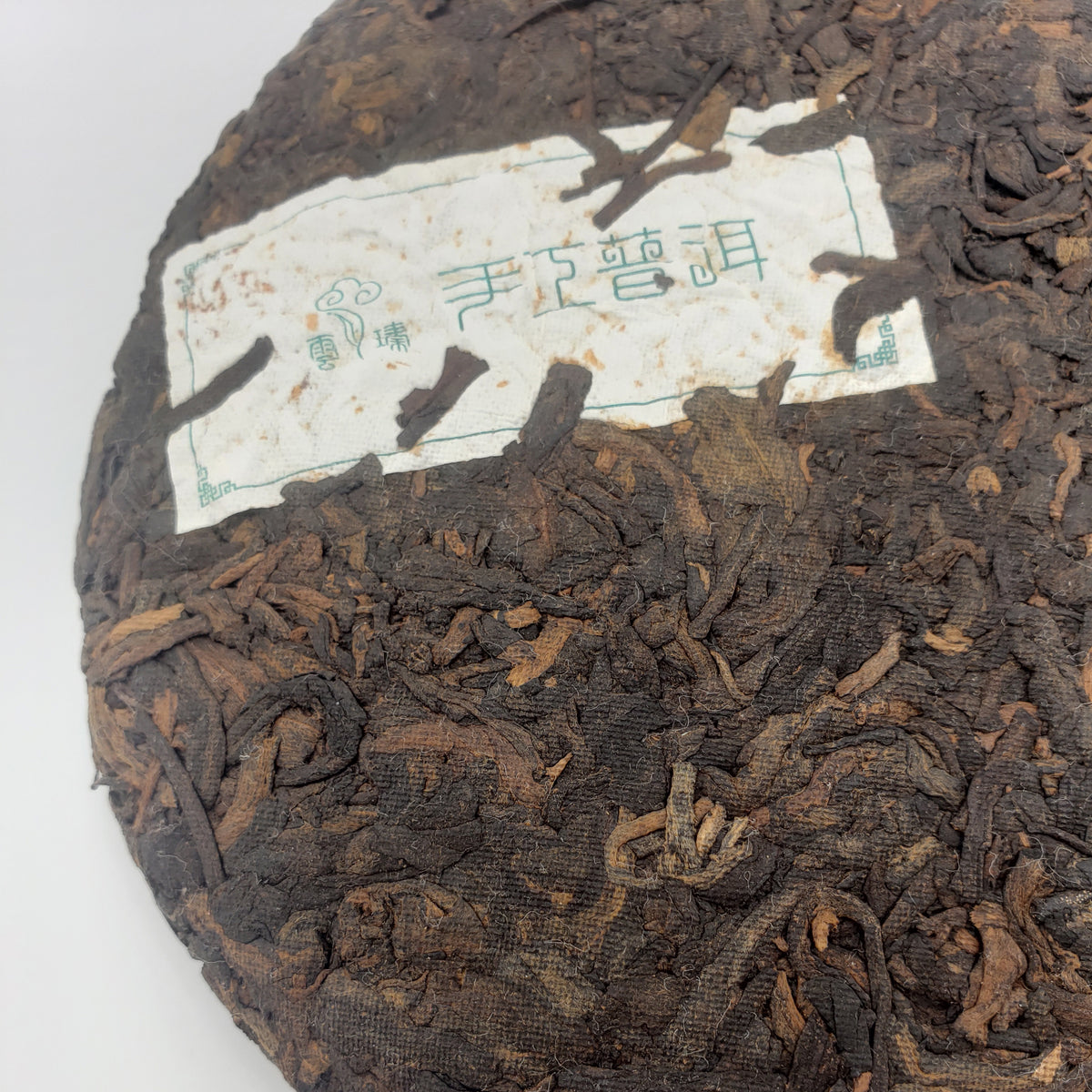A close-up of a round compressed tea cake with visible loose tea leaves and a white paper label. The label has green Chinese characters and a small logo, but the brand text is not clearly legible in Latin script, so no visible brand is listed.