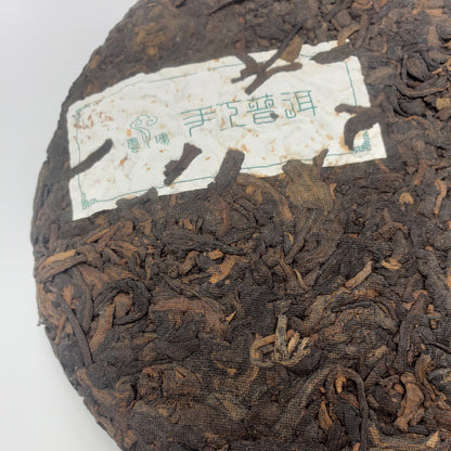 A close-up of a round compressed tea cake with visible loose tea leaves and a white paper label. The label has green Chinese characters and a small logo, but the brand text is not clearly legible in Latin script, so no visible brand is listed.