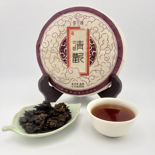 A round cake of Chinese tea on a small stand, with a cup of brewed tea and a dish of loose leaves in front, photographed on a clean white background.