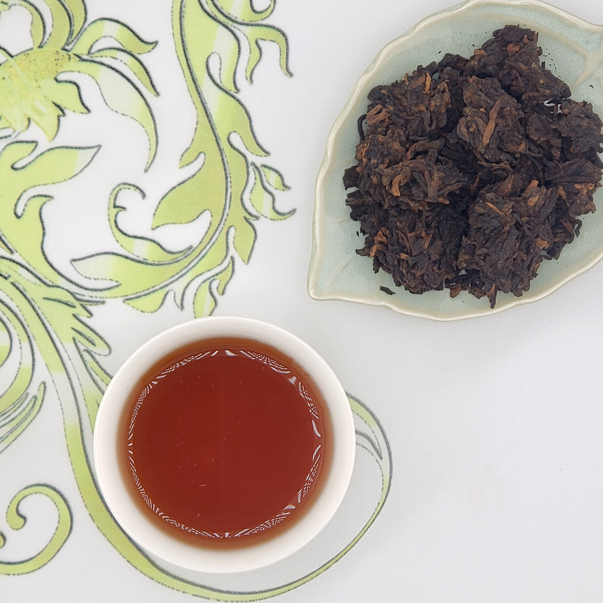 A white teacup filled with amber-brown tea sits beside a small dish of dark dried tea leaves on a white surface with a light green ornamental pattern.