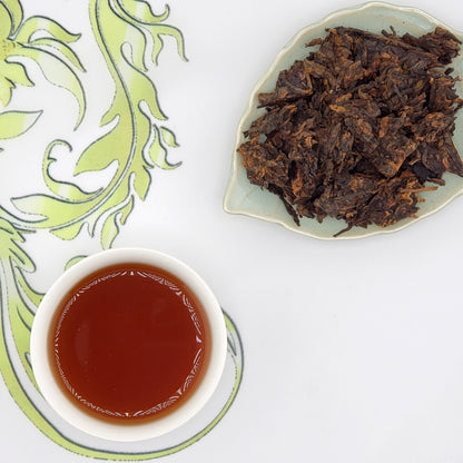 Dried tea leaves on a plate with a cup of brewed tea on a decorative mat.
