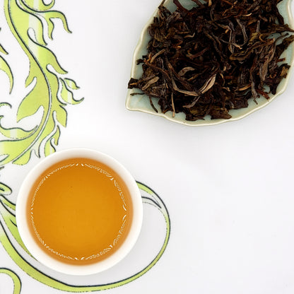 Dry tea leaves on a leaf-shaped plate with a cup of brewed tea on a decorative mat.