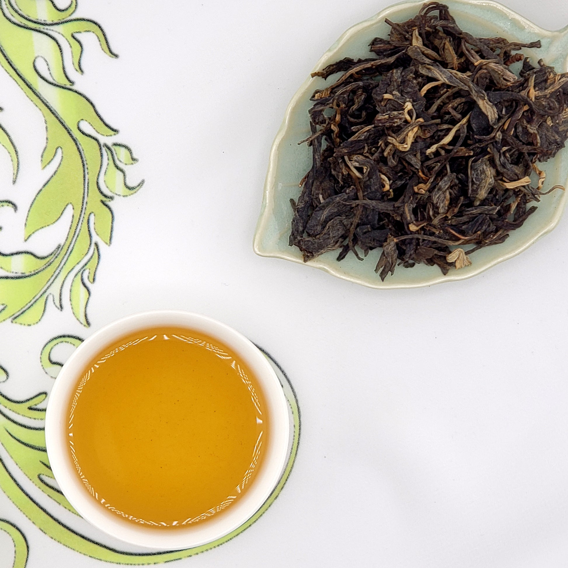 Dry tea leaves on a leaf-shaped dish with a cup of brewed tea on a decorative saucer.
