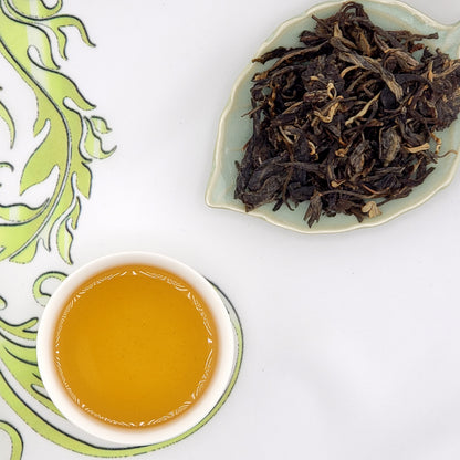 Dry tea leaves on a leaf-shaped dish with a cup of brewed tea on a decorative saucer.