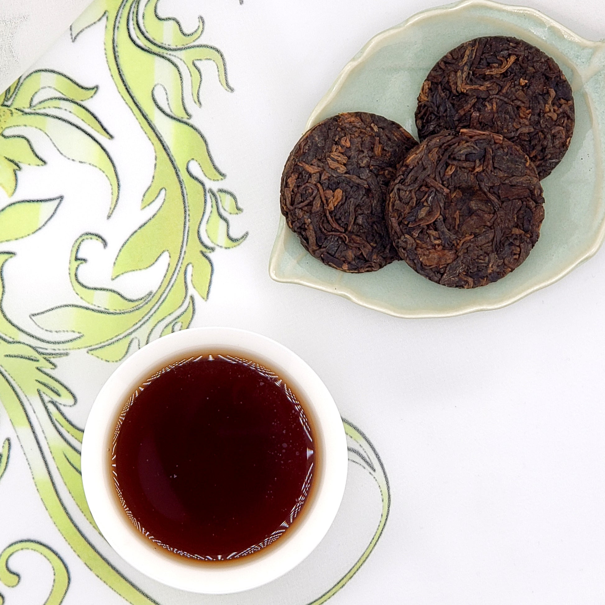 Three dark brown pressed tea coins with a small cup of brewed tea on a decorative white and green background.