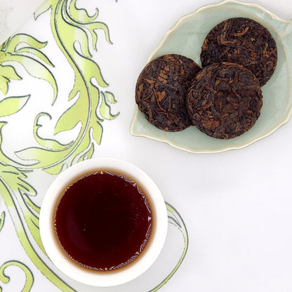 Three dark brown pressed tea coins with a small cup of brewed tea on a decorative white and green background.