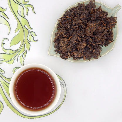 Dried tea leaves on a leaf-shaped plate with a cup of brewed tea on a decorative mat.